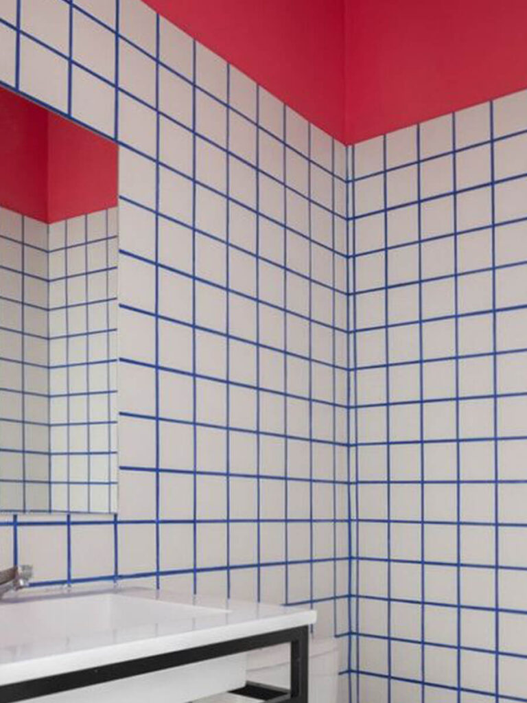 Bathroom walls covered in white tiles with blue grout, topped by a vivid pink-red painted upper wall and ceiling, reflected in a large mirror.