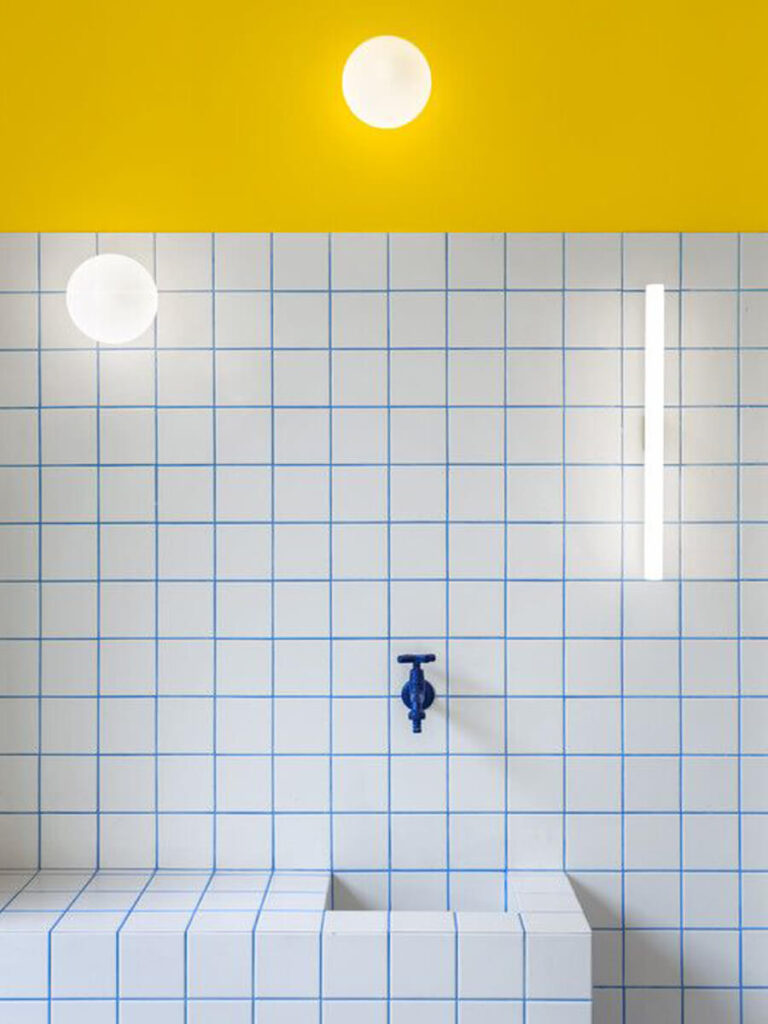 Bathroom with white square tiles and vivid blue grout, bold yellow wall and ceiling above the tiles, and globe light fixtures for a playful, modern look.