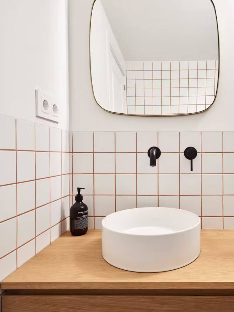 Minimalist bathroom with white tiles and brown grout, light wood counter, black faucet, and round white basin for contrast.