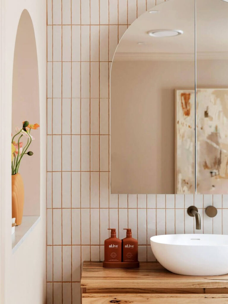 Contemporary vanity with vertical white tiles and warm brown grout, wooden counter, curved mirror, and peach-beige painted walls for a soft aesthetic.