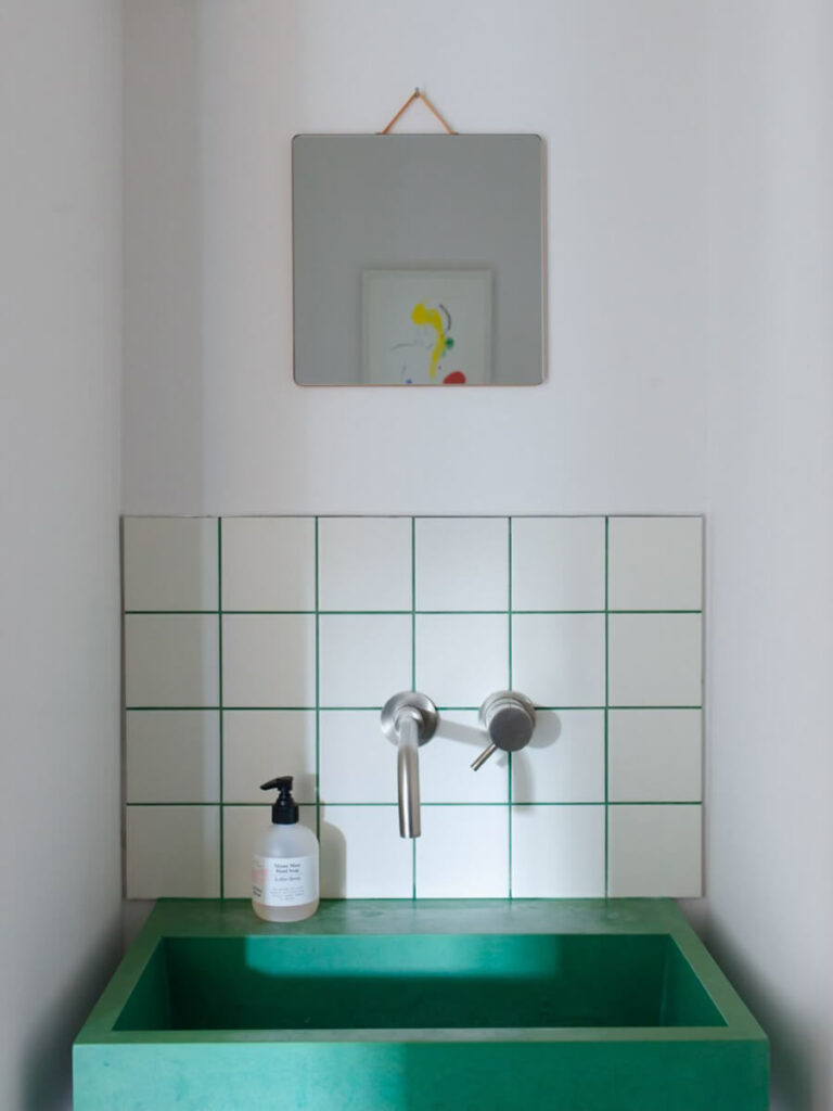 Minimal bathroom sink area with white tiles featuring green grout lines, an emerald green basin, wall-mounted faucet, and square mirror above.