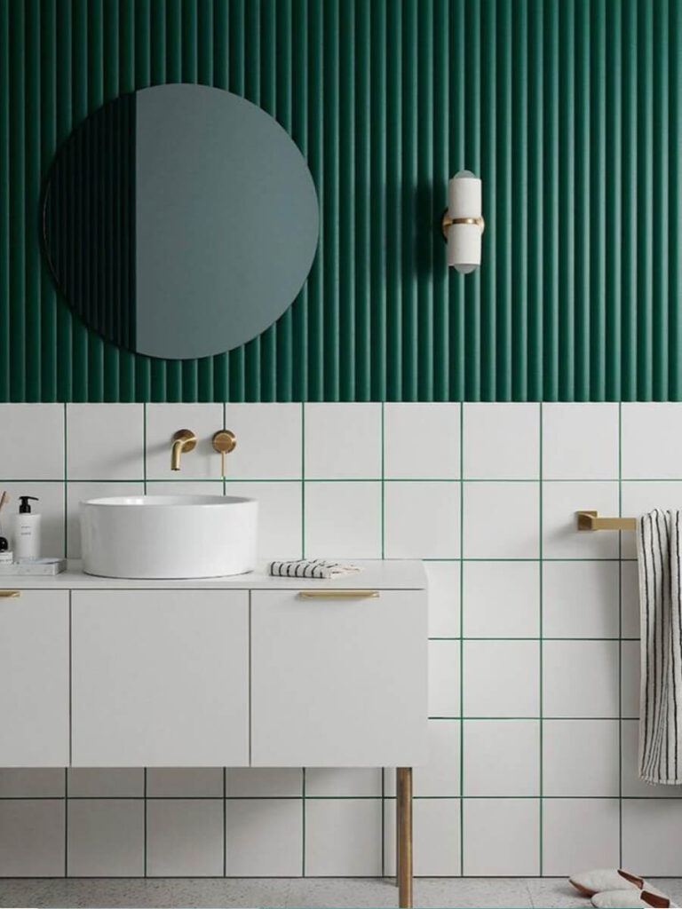 Modern bathroom vanity with white tiles and green grout, deep green vertical panel wall, round mirror, and brass fixtures.