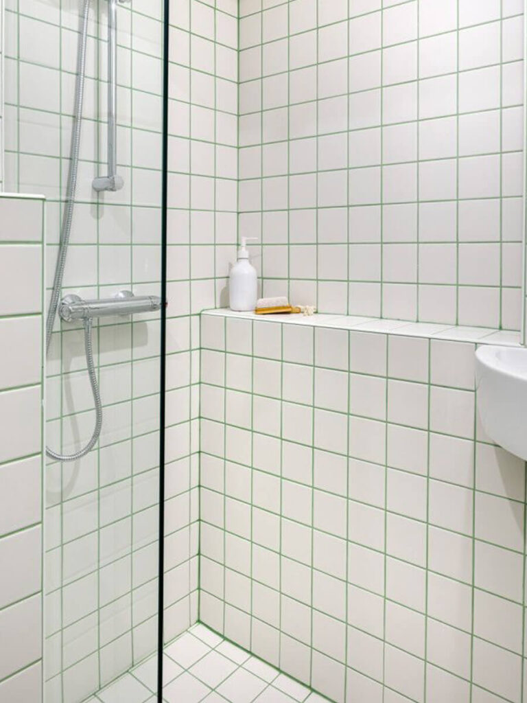 Shower area with white tiles and green grout, glass partition, built-in shelf with accessories, and chrome fixtures for a crisp, fresh look.