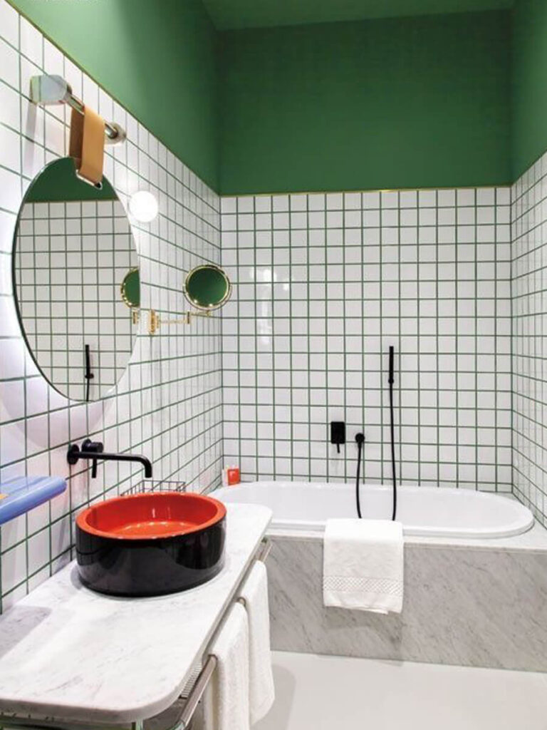 Eclectic bathroom with white tiles and green grout lines, green-painted ceiling and walls, marble vanity, and round mirror along with a bold red basin.