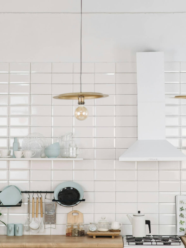 Kitchen backsplash of glossy white tiles with neutral gray grout, open shelves, blue and white ceramics, and modern pendant lighting above wood countertop.