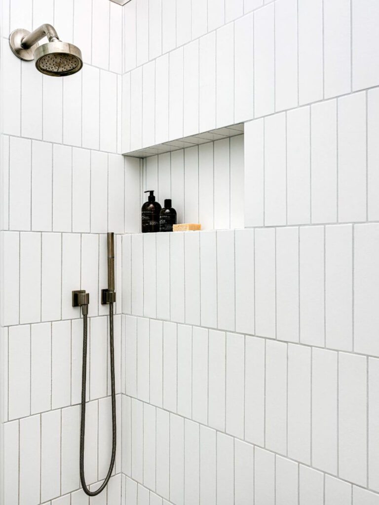 Minimalist shower with vertical white tiles and neutral gray grout, built-in shelf with toiletries, steel shower fixture, and clean lines throughout.