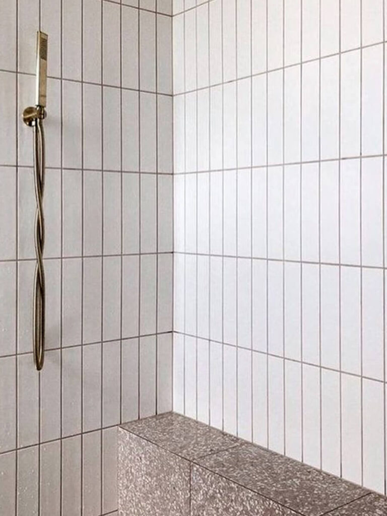 Shower with tall vertical white tiles, gray grout outlines, terrazzo bench, brass fixtures, and minimalist styling.