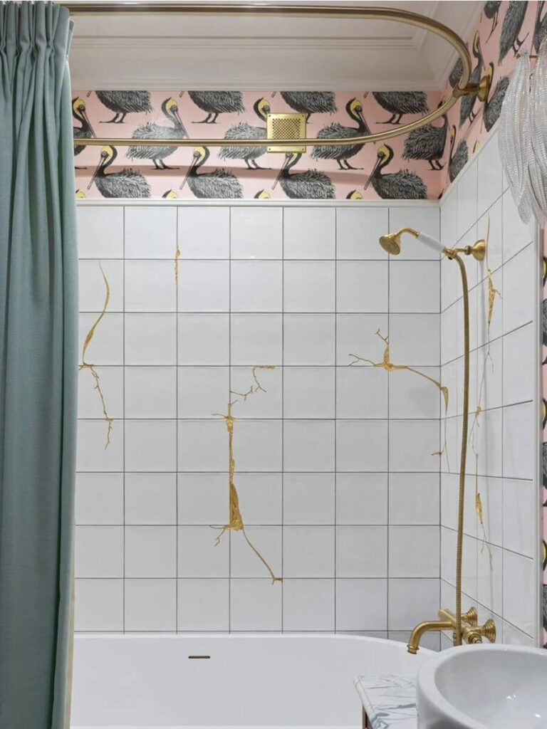 Bathroom with glossy white square tiles featuring gold metallic grout in a kintsugi-inspired pattern, brass fixtures, green curtain, and pelican-themed pink wallpaper.