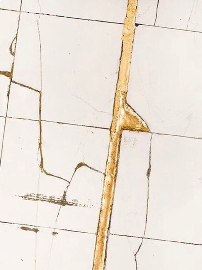 Close-up of glossy white tile with metallic gold grout lines, showing kintsugi-repaired cracks for artistic bathroom design.