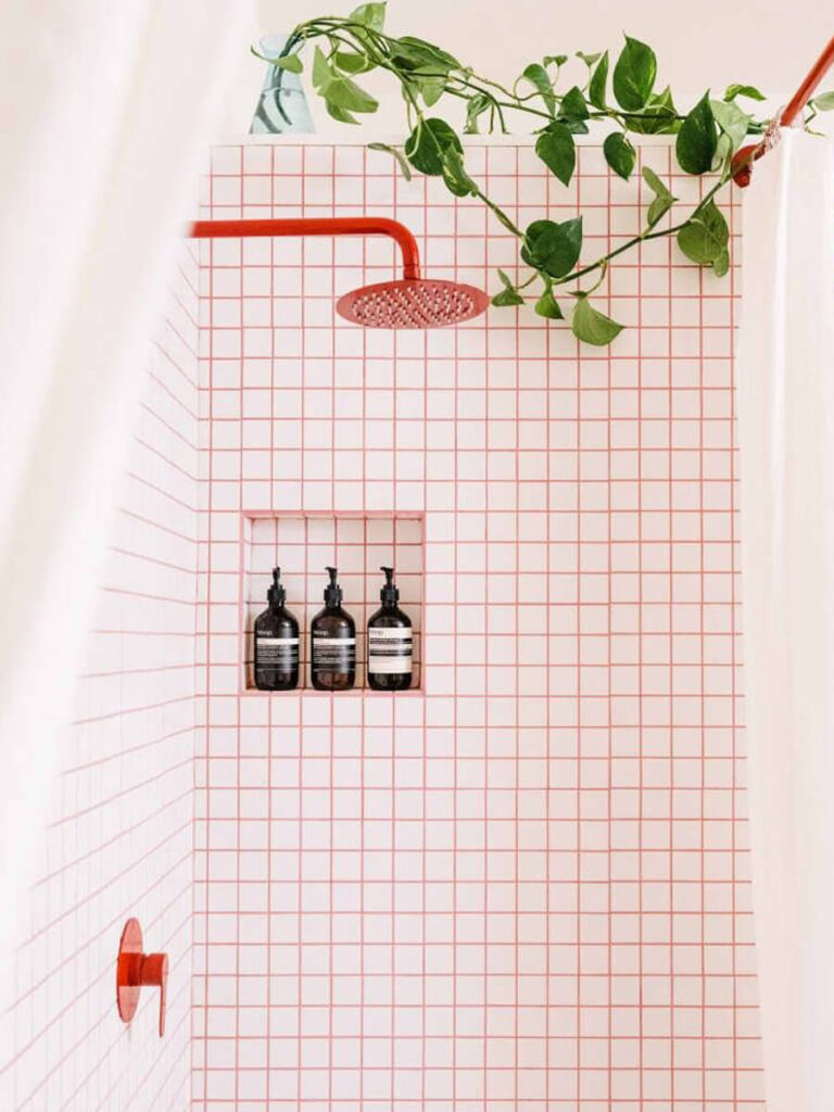 Bathroom shower corner with square white tiles and bold orange grout, matching orange fixtures and lush green trailing plant.