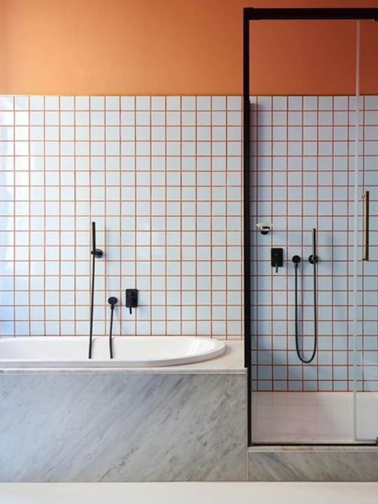 Bathroom with white square tiles, vivid orange grout, matching orange wall paint, marble bath surround, and matte black fixtures.