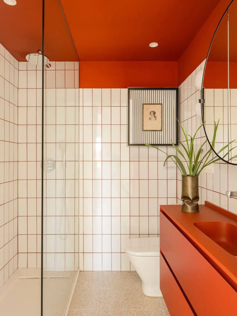 Bathroom with vertical white tiles, outlined with orange grout, bright orange ceiling and vanity, terrazzo flooring, and graphic wall art.