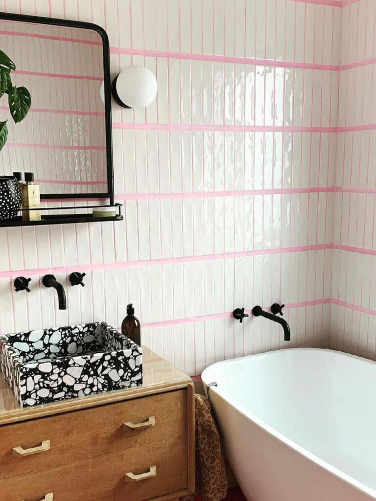 Bathroom with glossy white vertical tiles outlined in pink grout, black fixtures, wood vanity, and a terrazzo-patterned sink for a bold, contemporary style.