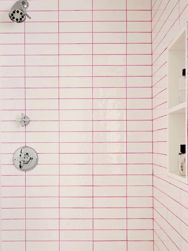 Shower with elongated white tiles in a grid pattern, outlined in pink grout, with chrome fixtures and cubby shelves for accessories.