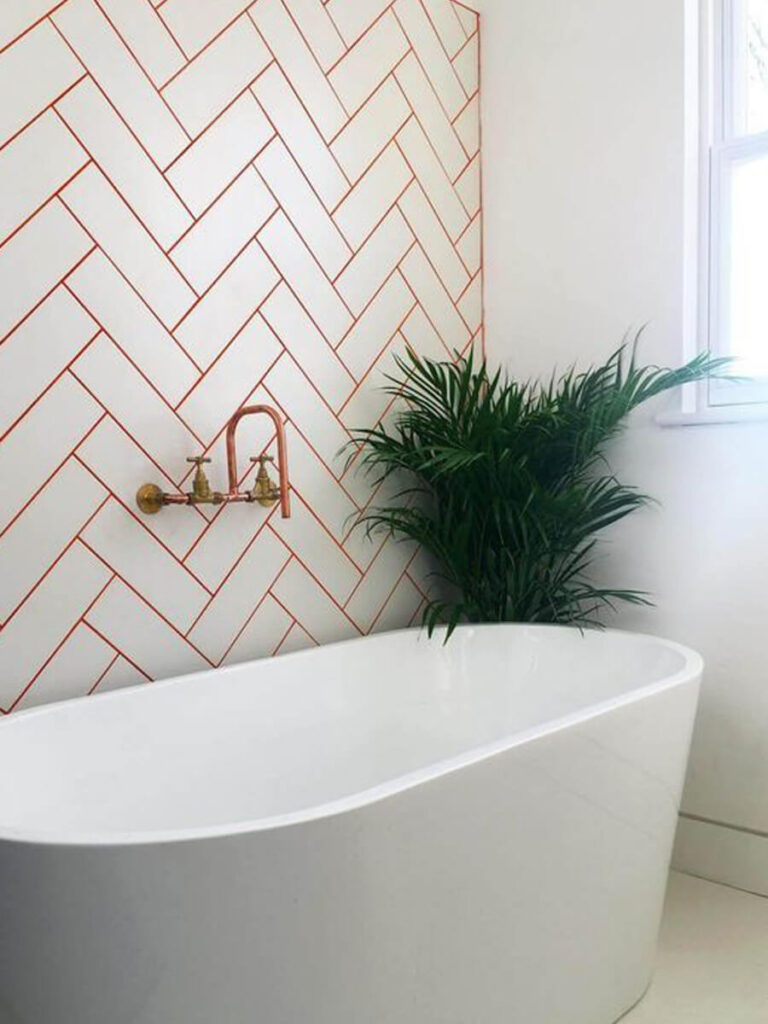 Bathroom with white herringbone wall tile, vivid red grout, gold fixtures, and lush plants for lively contrast.