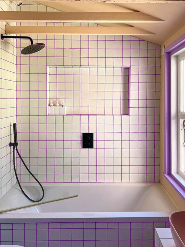 Bathtub alcove featuring white square tiles with violet grout, black shower hardware, light beige paint, wood ceiling beams, and purple window trim for a playful and modern look.