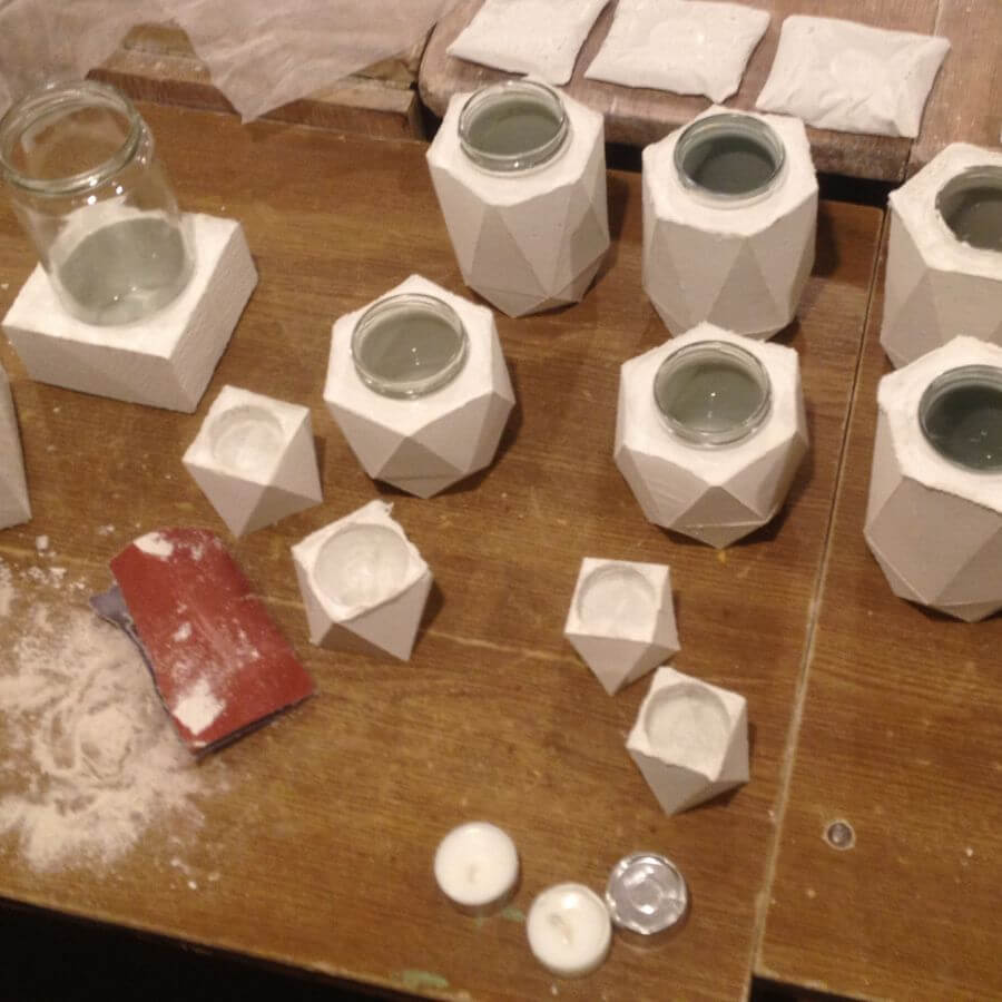 White cement diy craft geometric candle holders and small tealight cups arranged on a wooden table with sanding tools and a glass jar.