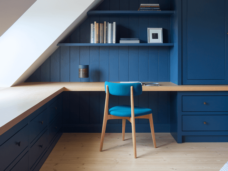Small attic bedroom with low ceiling and angled white walls, featuring navy blue built-in desk, shelving, and a bright workspace for creative attic bedroom designs.