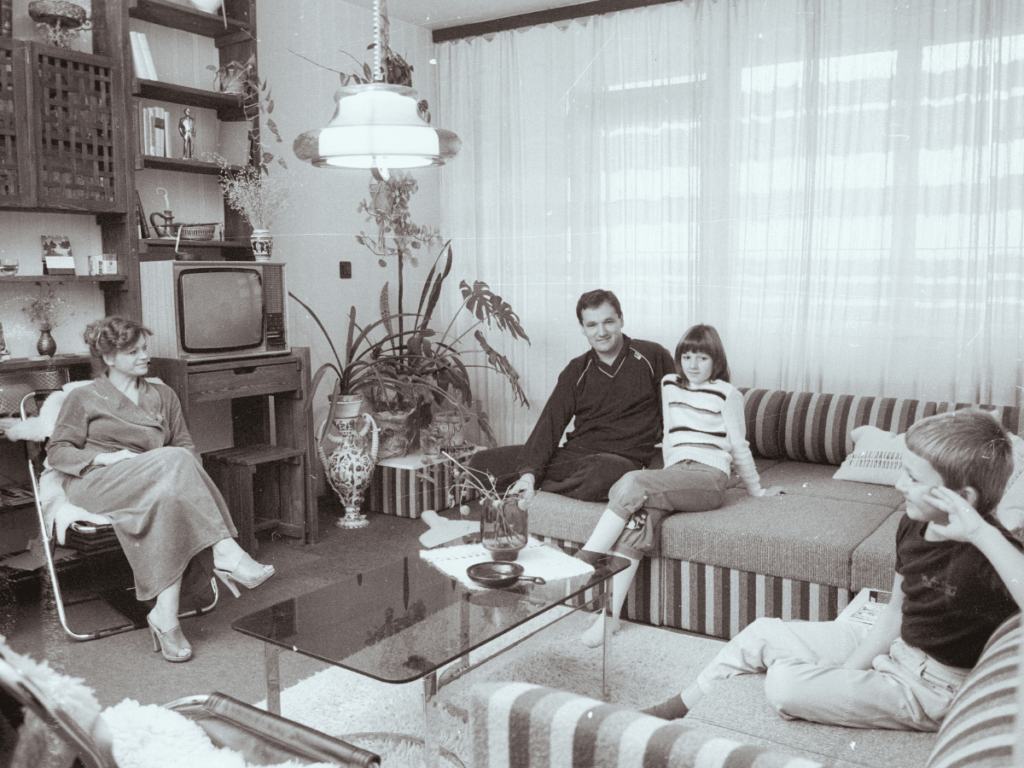 A family sitting on couches in a small living room. black and white foto from 1981, large window and plants, a square coffee table.