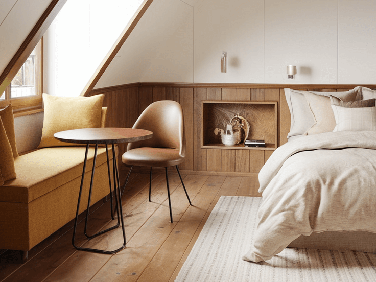 Small attic bedroom with slanted ceiling, built-in bench, round table, warm wood accents, and neutral cozy bedding.